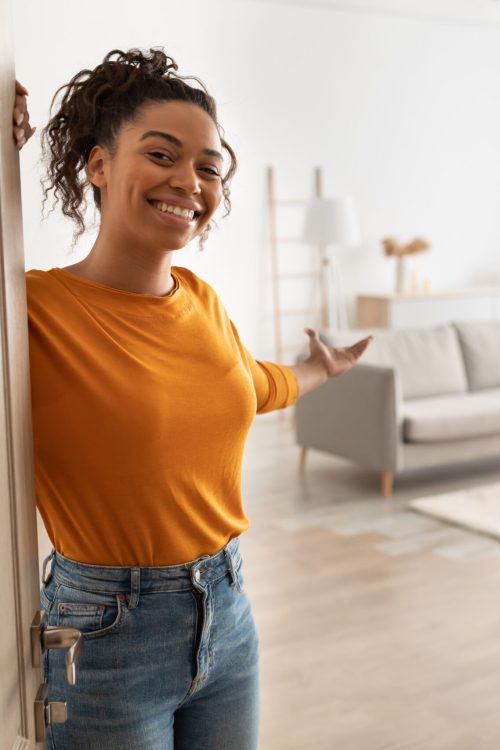 Cheerful African American Woman Opening Door And Gesturing Welcoming You To Come In Smiling To Camera Standing At Home. Hospitality, Real Estate Ownership And Purchase Concept